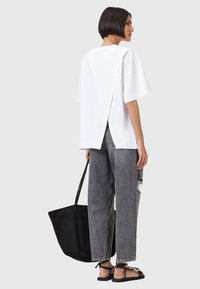 Witte oversized t-shirt met een gespleten achterkant, gecombineerd met grijze versleten jeans en platte zwarte sandalen. Zwarte tote bag wordt in één hand vastgehouden.