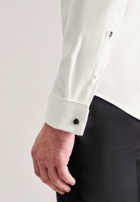 Camicia bianca a maniche lunghe con un bottone nero al polsino, con un piccolo motivo floreale ricamato in nero vicino alla manica. Texture liscia.