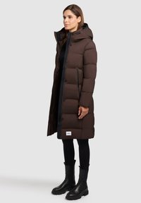 Giacca imbottita marrone dal design lungo, con cerniere laterali e cappuccio nero. Tessuto strutturato con sezioni trapuntate visibili e etichetta con logo. Pantaloni e stivali neri.