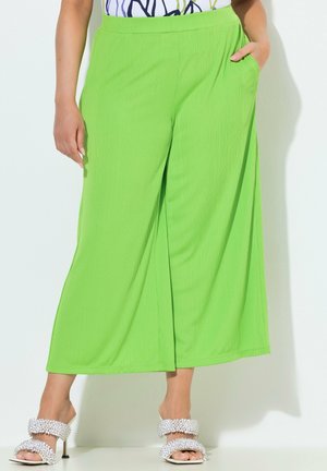 Frau trägt leuchtend grüne, weit geschnittene Hosen und weiße Sandalen mit Absatz und strukturierten Riemen, stehend vor weißem Hintergrund.