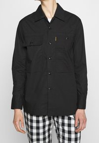 Camicia nera con bottoni e due tasche frontali, dotata di colletto a punta e texture uniforme. Abbinata a pantaloni a scacchi bianchi e neri.