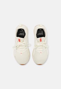 Białe buty sportowe z teksturowanym siatkowym wierzchem, czarnymi wkładkami, jasnymi sznurówkami i czerwonym akcentem na języku. Gumowe podeszwy zapewniające przyczepność.