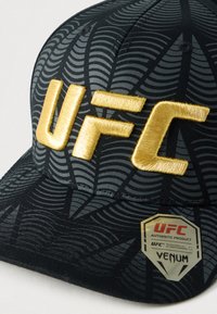 Μαύρο καπέλο με ανάγλυφο, σχεδιασμένο ύφασμα. Διαθέτει έντονα, χρυσά γράμματα "UFC" και ετικέτα στο πλάι που υποδεικνύει την αυθεντικότητα και το εμπορικό σήμα.