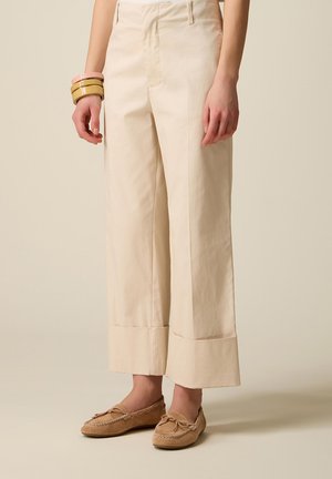 Pantaloni beige a taglio dritto con ampi risvolti, indossati con mocassini marrone chiaro e braccialetti impilati al polso sinistro.