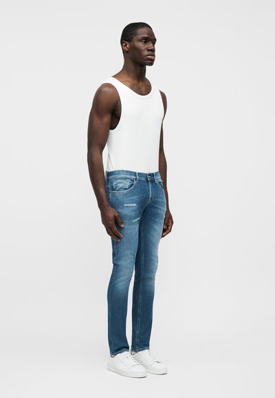 Tenue masculine comprenant un débardeur blanc et un jean skinny bleu clair déchiré, complétée par des baskets blanches, sur un fond neutre.