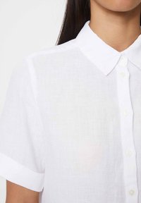 Camicia bianca in lino con colletto classico, maniche corte arrotolate e chiusura frontale con bottoni bianchi e tessuto testurizzato.