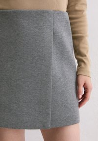 Grauer Baumwollrock mit glatter Textur und einer frontal verlaufenden Naht. Kombiniert mit einem teilweise sichtbaren, sandfarbenen Langarmshirt.