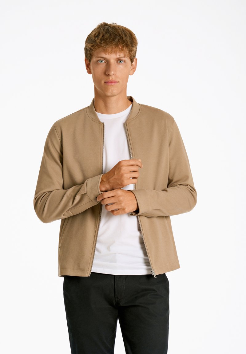 Chaqueta cremosa con cremallera y cuello alto, mangas largas y un diseño ajustado, superpuesta a una camiseta blanca. Llevada con pantalones negros.