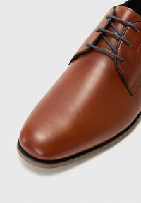 Brown leather shoe with a smooth finish, rounded toe, and contrasting gray laces. Features a rubber sole with a slight textured edge.