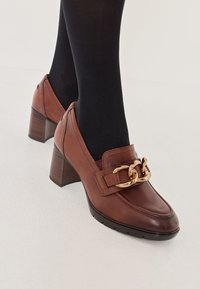 Scarpe con tacco cubano in pelle marrone, con punta arrotondata, presentano un accento in catena dorata sul collo del piede e dettagli strutturati sul tacco.