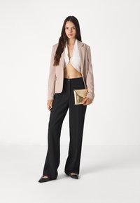 Beige blazer, white top, black wide-leg trousers, and black pointed flats. Holding a gold envelope clutch. Smooth textures and tailored fit.