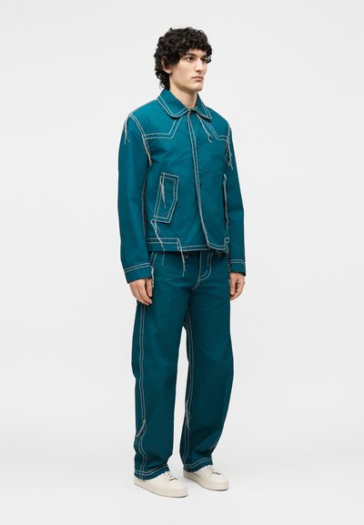 Νεαρός άνδρας που φοράει σακάκι και παντελόνι σε πετρόλ μπλε με λευκές αντίθετες ραφές και ξεφτισμένα άκρα, συνδυασμένα με λευκά αθλητικά παπούτσια, στέκεται μπροστά σε λευκό φόντο.