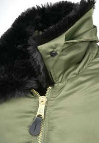 Alyvuogių žalia striukė su juodu dirbtinio kailio apykakle, auksiniu užtrauktuku su juodu traukimo galiuku ir audiniu, turinčiu lygų, tekstūruotą paviršių.