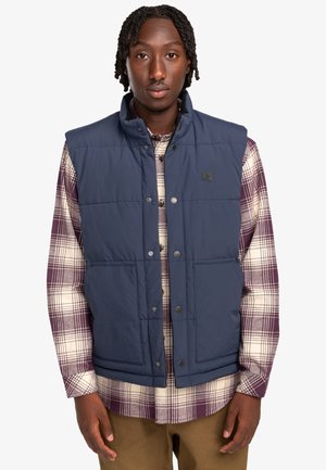 Navy blue puffer vest with a high collar, snap-button closure, and two front pockets, worn over a plaid shirt. Textured fabric.