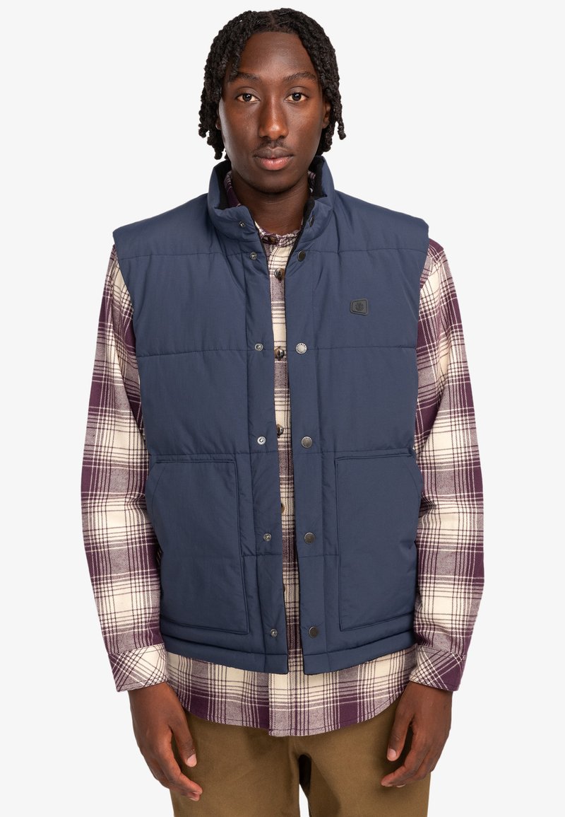 Navy blue puffer vest with a high collar, snap-button closure, and two front pockets, worn over a plaid shirt. Textured fabric.