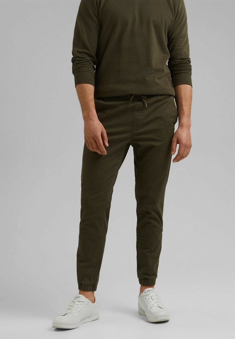 Homme portant un pantalon de jogging vert foncé, un haut à manches longues assorti et des baskets blanches Esprit, debout devant un fond uni.