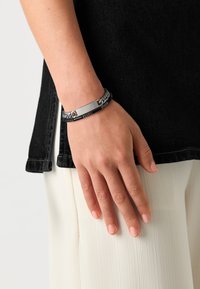 Pulsera de metal y tejida en tonos plateados, con un centro plano y rectangular y acentos texturizados, llevada en una mano de piel clara.