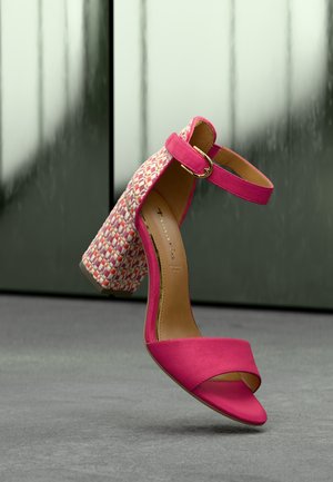 Pink suede high-heeled sandal with ankle strap and multicolored woven block heel, shown against a blurred green and gray background.