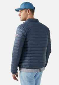 Giubbotto puffer blu navy, design trapuntato con cuciture orizzontali, colletto rialzato e patch con logo sulla manica sinistra. Indossato sopra una maglietta grigia.