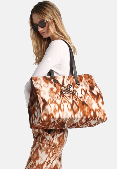 Woman wearing sunglasses and white top carries large brown and white patterned tote bag with black handles over shoulder.
