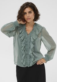 Blouse transparente vert clair avec des détails volantés à l'avant et au col, dotée de manches longues avec des poignets froncés et d'un décolleté en V.