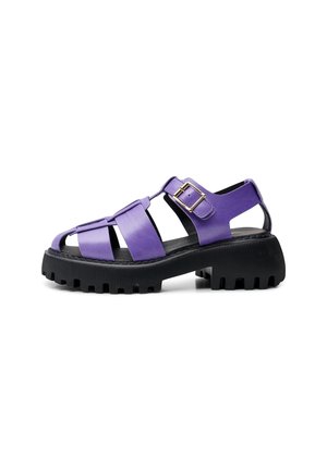 Lilla lædersandal med kraftig sort ridsesål, flere remme og gyldent spænde på siden.