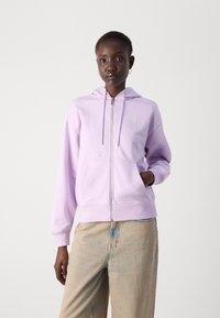 Šviesiai violetinė zip-up džemperis su priekinėmis kišenėmis, su raišteliais kapuci, ir logotipu ant krūtinės, derinamas su šviesiomis džinsinėmis kelnėmis.
