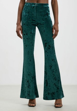 Pantaloni a zampa d'elefante in velluto di un profondo teal con una texture liscia, caratterizzati da una vita a mezza altezza e un accento circolare sulla parte frontale.