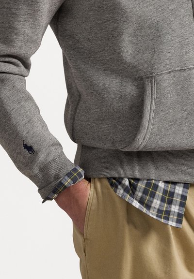 Hanorac gri cu manșete și tiv cu nervuri, cu un logo brodat pe mânecă, suprapus peste o cămașă în carouri și pantaloni khaki.