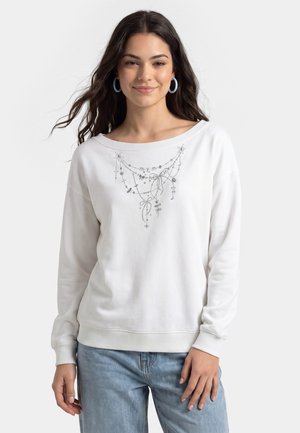 Ung kvinde iført hvid sweatshirt med delikat sølvkæde og sløjfedetalje, kombineret med lyseblå jeans og blå creoler.