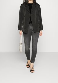 Mujer con blazer negro de doble botonadura, top negro, jeans ajustados grises, tacones negros de tiras, sosteniendo un pequeño bolso blanco, de pie sobre un suelo liso.