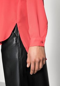 Korallenfarbige, durchsichtige Bluse mit langen Ärmeln, abgerundetem Saum und geknöpften Manschetten, kombiniert mit einem eleganten, schwarzen Lederrock.