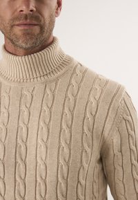 Beżowy sweter z golfem o splocie w warkocze, z prążkowanym kołnierzem, teksturowanymi wzorami i dopasowanym krojem, wykonany z miękkiego materiału.