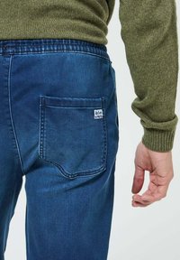 Pantalon en denim bleu à coupe décontractée, avec une poche arrière et une taille élastique. Le tissu a une texture lisse et peu de décoloration.
