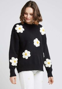 Pull noir en tricot avec des appliqués de marguerites blanches texturées et des centres jaunes, col rond et manches longues à côtes.