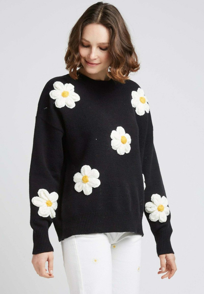 Pull noir en tricot avec des appliqués de marguerites blanches texturées et des centres jaunes, col rond et manches longues à côtes.