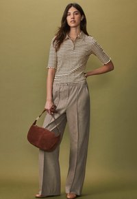 Polo shirt beige à rayures avec manches courtes, associé à un pantalon ample gris clair et un sac à main en daim marron. Fond de couleur unie.