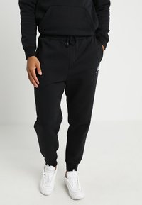 Černé tepláky s šňůrkou v pase a bočními kapsami, na levém stehně s bílým logem. Nosené s bílými teniskami.