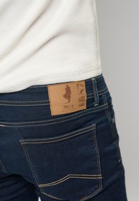 Tamno plavi traperice s kožnom etiketom koja ima siluetu i logo branda. Vidljivi su detaljni šavovi i dizajn džepova.