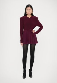 Μακρυμάνικο φόρεμα σε βαθύ βουργουνδί χρώμα με φουσκωτή φούστα, ζώνη στη μέση και V-λαιμόκοψη. Φορεμένο με μαύρο καλσόν και μυτερά τακούνια.