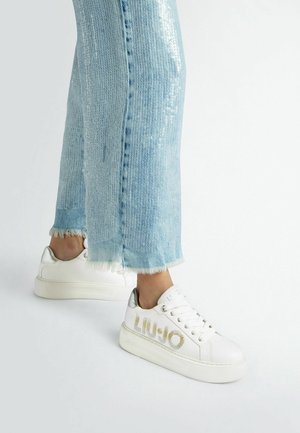 Sneakers in pelle bianca con accenti argento e suola testurizzata, abbinati a jeans a zampa leggera di colore blu chiaro con orli sfrangiati e motivi di paillettes.