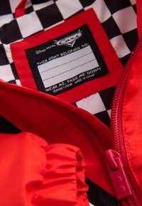 Rote Jacke mit pinkem Reißverschluss und kariertem schwarz-weißem Innenfutter, das ein Etikett mit der Aufschrift "This coat belongs to" und dem Disney Pixar Cars-Logo zeigt.