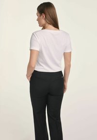 Kobieta z brązowymi włosami, ubrana w białą koszulę z krótkim rękawem i czarne spodnie, stojąca z rękami w kieszeniach, odwrócona plecami w stosunku do jednolitego tła.