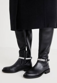 Les bottes en cuir noires montantes jusqu'au genou présentent une fermeture éclair, des sangles ornées d'accents argentés et un talon bloc pour la stabilité et le confort.