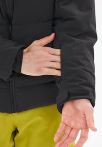 Svart puffjacka med dragkedja och kardborrestängning. Har en slät textur och en enkel, strömlinjeformad design.