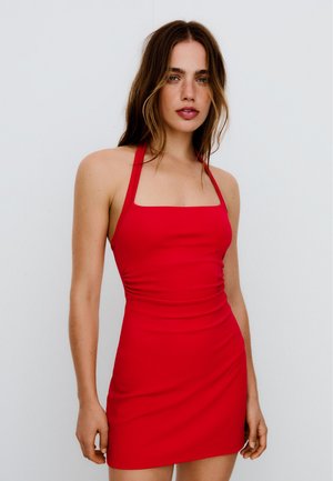 Mladá žena s voľne rozloženými hnedými vlasmi, oblečená v priliehavých červených mini šatách s halter výstrihom, stojaca pred jednoduchým bielym pozadím.