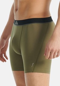 Detailní záběr muže v olivově zelených těsných boxerách s černým pasem a značením Climacool na stehně.