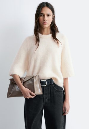 Kvinde med langt mørkt hår iført en hvid, blød sweater og sorte bukser med bælte, holder en stor taupe clutch taske mod en ensfarvet lys baggrund.