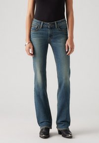 Femme portant un jean bootcut bleu délavé, un top noir, un bracelet et des bagues en argent, ainsi que des bottes noires à bout pointu, debout devant un fond uni.