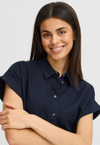 Cămașă bleu marin cu mânecă scurtă, cu guler pliant și detaliu cu nasturi, realizată dintr-un material texturat. Modelul stă cu brațele încrucișate.